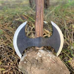 May include: A double-bladed axe with a dark gray head and a wooden handle. The blades have a curved shape with a silver edge. The handle is made of brown wood. The axe is resting on a rock.