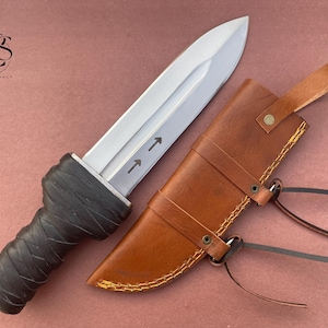 May include: A silver dagger with a black handle and a brown leather sheath with stitching. The dagger has two arrows pointing upwards on the blade.