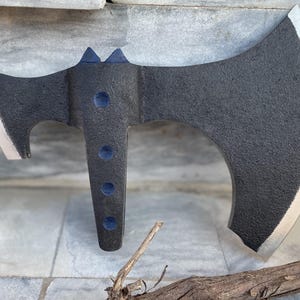 Könnte beinhalten: Eine schwarze Axt mit strukturierter Oberfläche und silberner Klinge. Die Axt hat eine gebogene Klinge, einen Haken und blaue Akzente. Die Axt liegt auf einer Steinoberfläche.