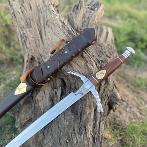May include: A silver and wood sword with a leather sheath. The sword has a twisted silver handle and a wooden grip with a gold emblem. The sheath is brown leather with a strap.