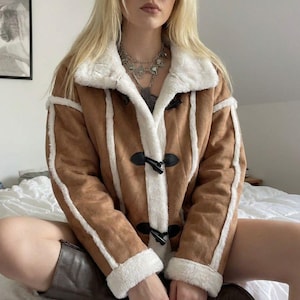 May include: A brown suede jacket with white faux fur trim and accents. The jacket has a toggle closure and is worn with brown boots. The jacket has a classic, vintage style.