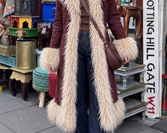 May include: A long, dark burgundy coat with beige faux fur trim on the collar, cuffs, and hem, worn over a brown top and dark blue flared jeans. The outfit includes a beige wide-brimmed hat, a brown crossbody bag, and dark brown platform shoes.