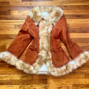 May include: A burnt orange suede jacket featuring a thick, cream-colored faux fur trim along the collar, front opening, and sleeve cuffs. The jacket includes two front patch pockets and a visible white faux fur lining throughout the interior.