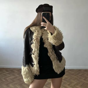 Könnte beinhalten: Eine dunkelbraune Lederjacke mit dickem, cremefarbenem, lockigem Lammfellbesatz an der Vorderseite, am Kragen und an den Ärmelbündchen. Die Jacke verfügt über Knebelverschlüsse und wird mit einem dunkelbraunen Barett und einem schwarzen Kleid kombiniert.