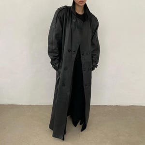 May include: A long, black leather trench coat featuring a double-breasted button closure, a high collar, structured shoulders with epaulets, and long sleeves with buttoned cuffs. The coat extends to the lower calf and is worn over black clothing.