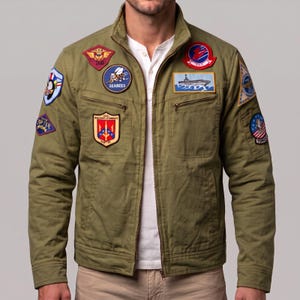 May include: An olive green utility jacket featuring a stand-up collar and a full-length front zipper. The jacket is adorned with various embroidered military-style patches, including a Seabees bee, a naval aircraft carrier, and a Tomcat emblem.
