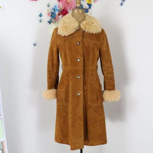 May include: A long, tan suede coat featuring a thick, cream-coloured faux fur collar and matching fur cuffs at the wrists. The coat has a button-front closure with five visible buttons, two front patch pockets, and visible stitching details.