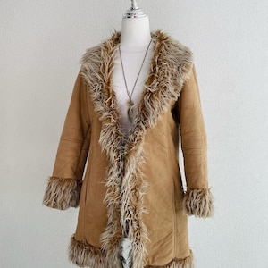 May include: A tan faux suede coat with faux fur trim on the collar, cuffs, and front edges. The coat is displayed on a mannequin. A long, flowing design. A silver necklace is visible.