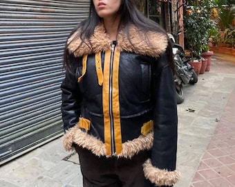May include: Black leather jacket with a tan faux fur collar and trim. The jacket features a gold zipper and accents, with gold straps on the shoulders and pockets. The jacket is worn over dark pants.