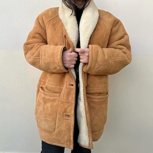 May include: A tan suede coat featuring a thick cream-colored shearling lining and a wide lapel collar. The coat includes long sleeves, two large front patch pockets, and a button-front closure. The exterior has visible seam detailing throughout.
