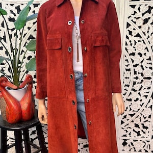 May include: A long, rust-colored suede coat featuring a classic collar, long sleeves, and a front button closure with metallic hardware. The coat includes two chest pockets and a straight hemline, worn over a white top and blue denim jeans.