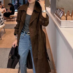 Könnte beinhalten: Ein langer, dunkelbrauner Wildleder-Trenchcoat mit breitem Kragen, langen Ärmeln mit Schnallenmanschetten und einem passenden Taillengürtel. Der Mantel wird über einem grauen Tanktop und einer hellblauen Jeans getragen, ergänzt durch eine schwarze Handtasche.