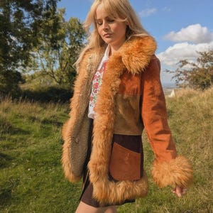 Puede incluir: Chaqueta de ante marrón de estilo vintage con cuello y puños de piel sintética. La chaqueta presenta un diseño de patchwork y un bolsillo grande en la parte delantera. La persona lleva una camisa blanca con diseño floral y una falda marrón.