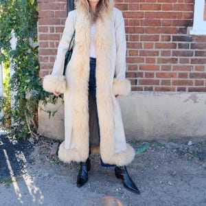 May include: A long, cream-colored coat with faux fur trim along the collar, front, and cuffs. The coat is worn with dark blue jeans and black pointed-toe boots. The background features a brick wall and a white picket fence.