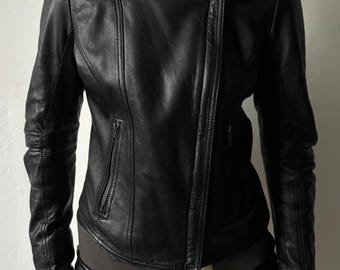 May include: A black leather jacket featuring a high collar, an asymmetrical front zipper closure, and two zippered pockets on the front. The jacket is paired with black leather shorts and a dark gray belt with a large, rounded metal buckle.