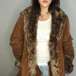 May include: A brown suede coat featuring a wide, plush faux fur collar and matching faux fur cuffs. The coat has visible light-coloured stitching along the sleeves and body, and is worn over a simple white top. The design includes long sleeves.