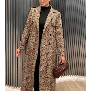 May include: A long trench coat featuring a tan and brown snakeskin print pattern, a notched collar, long sleeves with cuffs, and a double-breasted button closure. The coat is worn over a black mock neck top and paired with a dark brown handbag.