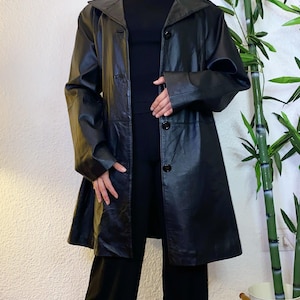 May include: A black leather coat featuring a wide collar, long sleeves, and a front button closure. The coat has a smooth finish and a straight hemline that falls below the hips. It is worn over a plain black top and black trousers.