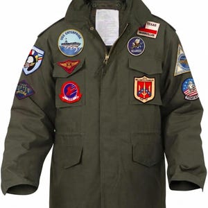 May include: An olive green field jacket featuring a high collar, a front zipper closure, and four flap pockets. The jacket is adorned with various embroidered military-style patches, including USS Enterprise, Seabees, and Tomcat emblems on the chest and sleeves.