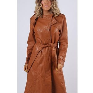 May include: A long, double-breasted trench coat in a rich cognac brown colour. The coat features a wide collar, shoulder epaulettes, long sleeves with buckled cuffs, and a matching tie belt at the waist. The smooth, structured material has a slight sheen.