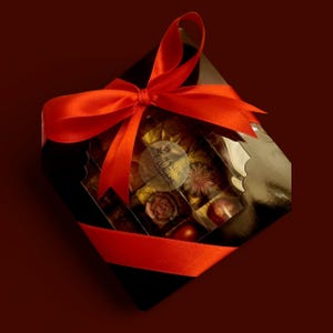 May include: A black, diamond-shaped gift box with a clear lid, filled with chocolates. The box is tied with a bright red satin ribbon. The chocolates are various shapes and colours, with a sticker that reads "MAISON CLUMIERE".