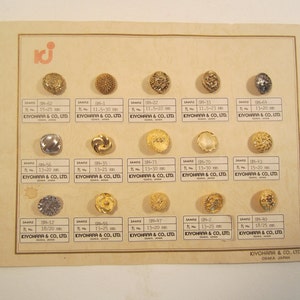 May include: A display card from Kiyohara & Co., Ltd. featuring fifteen assorted metal buttons in gold and silver tones. Each button is mounted above a label with a sample number and size range in millimeters, ranging from 11.5 to 30 mm.