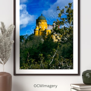 May include: A framed print featuring a yellow castle with a green dome, set against a blue sky with clouds. The castle is surrounded by green trees. The print is on a white wall above a wooden shelf. The text "OCMImagery" is visible.