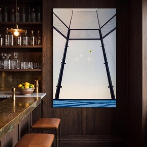Könnte beinhalten: Ein großes vertikales Wandbild, das einen Padel-Platz mit blauem Boden, schwarzem Metallzaun und einem einzelnen gelben Ball in der Luft vor einem hellen Himmel zeigt. Das Kunstwerk ist an einer dunkel getäfelten Holzwand montiert.