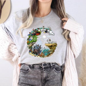 A woman wearing a gray t-shirt with a colorful graphic design, gray jeans, and a white cardigan stands in front of a wall with a hat and a potted plant.
