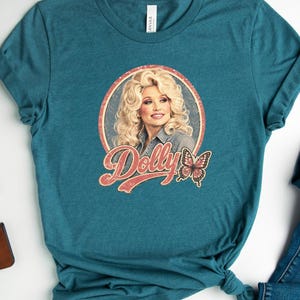 May include: A teal short-sleeved t-shirt featuring a graphic print of Dolly Parton inside a circular frame with the text Dolly and a butterfly illustration. The shirt is displayed alongside blue denim jeans and white canvas sneakers.
