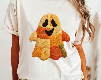 May include: A cream-colored t-shirt featuring a patchwork ghost design in shades of orange, yellow, and brown. The ghost has a smiling face with black eyes and mouth. The word "NATURAL" is printed in black at the bottom left.