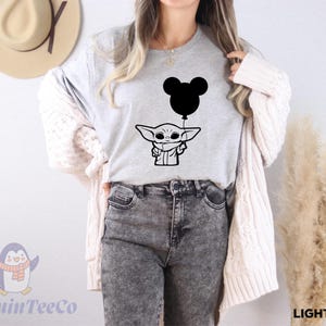 A woman wearing a gray t-shirt with a graphic of a cartoon character holding a balloon, standing in front of a wall with a hat and a potted plant.