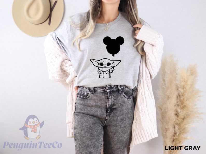 A woman wearing a gray t-shirt with a graphic of a cartoon character holding a balloon, standing in front of a wall with a hat and a potted plant.
