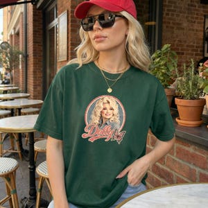 May include: A dark green short-sleeve t-shirt featuring a circular graphic of Dolly Parton with blonde hair, the text "Dolly" in a vintage red script font, and a small butterfly illustration. The shirt is worn with a red baseball cap and sunglasses.