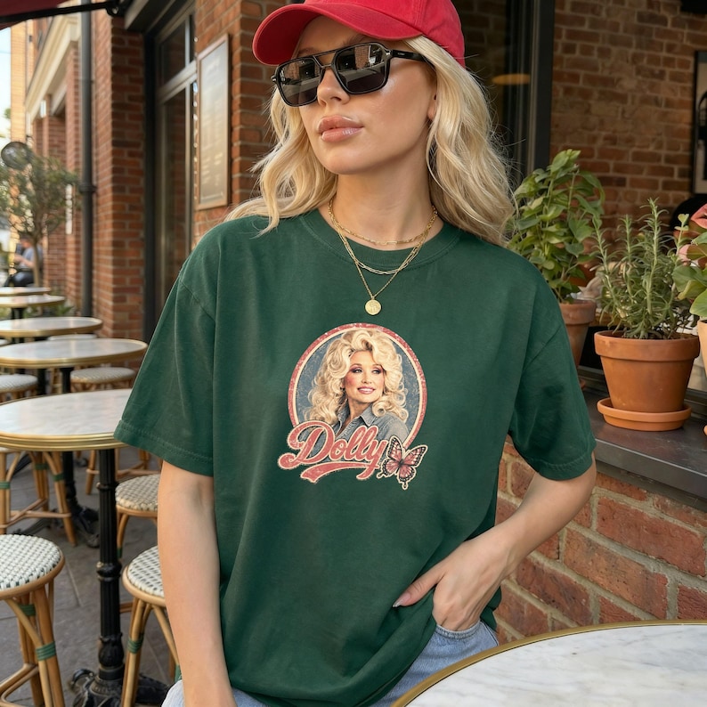 May include: A dark green short-sleeve t-shirt featuring a circular graphic of Dolly Parton with blonde hair, the text "Dolly" in a vintage red script font, and a small butterfly illustration. The shirt is worn with a red baseball cap and sunglasses.