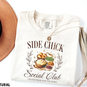 May include: Cream-colored t-shirt with the text "SIDE CHICK Social Club" and the phrase "I'M JUST HERE FOR THE SIDES". The graphic features illustrations of Thanksgiving side dishes. A gold necklace is draped over the shirt.