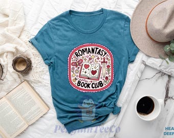 May include: A teal blue t-shirt with a pink and white graphic that says "Romantasy Book Club". The graphic features a book with hearts, a butterfly, a sword, and other whimsical details.