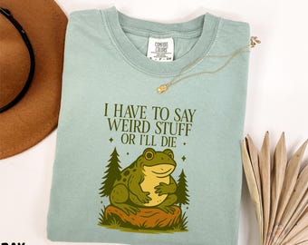 May include: A light blue t-shirt with the text "I HAVE TO SAY WEIRD STUFF OR I'LL DIE" and a cartoon frog illustration. The frog is green and yellow, sitting on a brown log with two green trees behind it. A gold necklace is draped on the shirt.