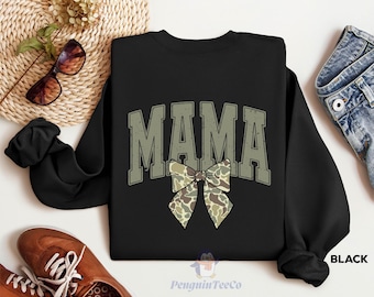 May include: Black sweatshirt with the word "MAMA" in green letters and a camouflage bow. The sweatshirt is on a white surface with a pair of brown shoes and a woven basket.