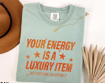 May include: A light green t-shirt with the text "YOUR ENERGY IS A LUXURY ITEM" in orange glitter. The shirt has a crew neck and short sleeves. A gold necklace with a pendant is draped on the shirt.