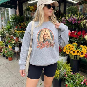 May include: A heather gray crewneck sweatshirt featuring a circular graphic of Dolly Parton with the text Dolly and a butterfly illustration. The sweatshirt has long sleeves and is paired with black athletic shorts and a beige baseball cap.