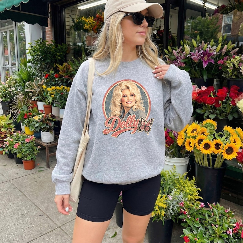 May include: A heather gray crewneck sweatshirt featuring a circular graphic of Dolly Parton with the text Dolly and a butterfly illustration. The sweatshirt has long sleeves and is paired with black athletic shorts and a beige baseball cap.