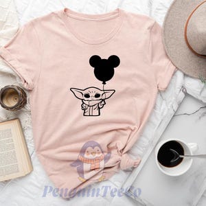 a pink t-shirt with a graphic of a baby alien character holding a balloon, along with various other items such as a hat, a cup, a book, and a clock.