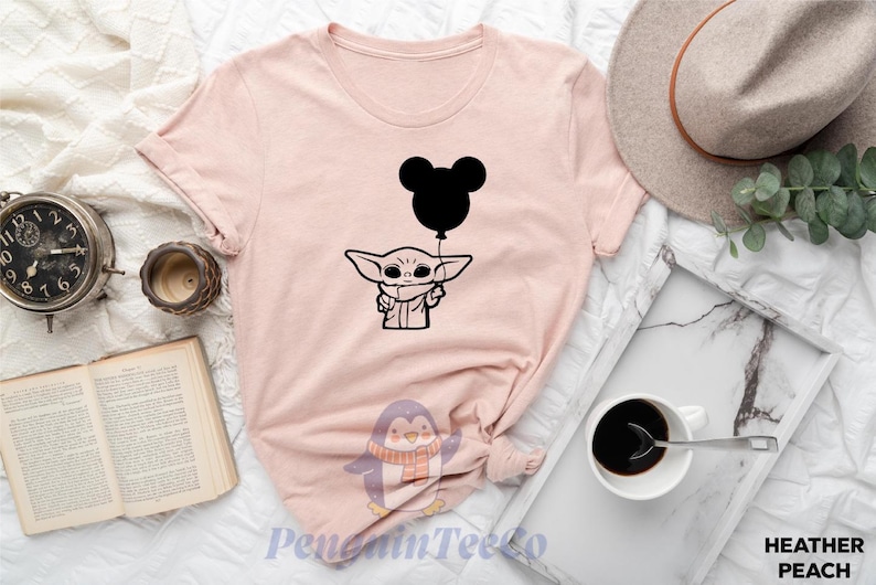 a pink t-shirt with a graphic of a baby alien character holding a balloon, along with various other items such as a hat, a cup, a book, and a clock.