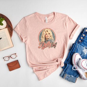 May include: A light pink short-sleeved t-shirt featuring a graphic print of a blonde woman with the text Dolly and a butterfly illustration. The shirt has a crew neckline and is displayed alongside blue denim jeans and white sneakers.