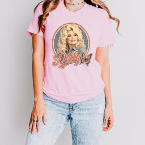 May include: A light pink short-sleeved t-shirt featuring a circular graphic print of a blonde woman with voluminous hair, the text "Dolly" in a retro-style font, and a small butterfly illustration. The shirt is paired with light blue denim jeans.