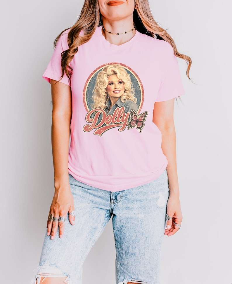 May include: A light pink short-sleeved t-shirt featuring a circular graphic print of a blonde woman with voluminous hair, the text "Dolly" in a retro-style font, and a small butterfly illustration. The shirt is paired with light blue denim jeans.