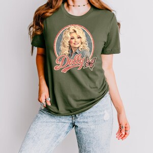 May include: An olive green short-sleeved t-shirt featuring a circular graphic of Dolly Parton with blonde hair, the text "Dolly" in a retro red font, and a butterfly illustration. The shirt is paired with light blue acid-wash denim jeans.