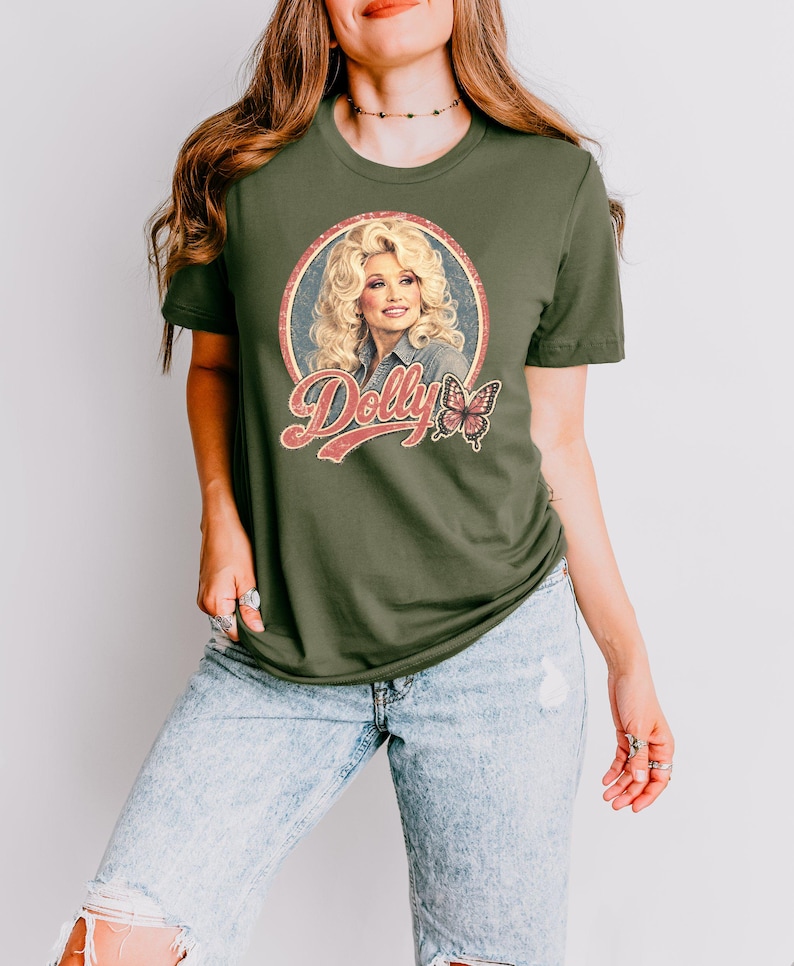 May include: An olive green short-sleeved t-shirt featuring a circular graphic of Dolly Parton with blonde hair, the text "Dolly" in a retro red font, and a butterfly illustration. The shirt is paired with light blue acid-wash denim jeans.