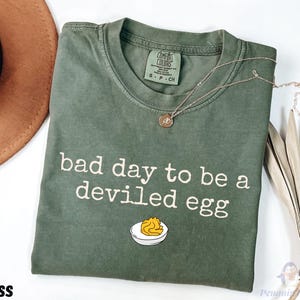 May include: Moss green t-shirt with the phrase "bad day to be a deviled egg" in white text. The shirt features a graphic of a deviled egg. A gold necklace and a brown hat are visible in the background.